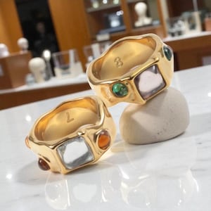 Op de afbeelding: Twee goudkleurige ringen met een getextureerde afwerking. Elke ring heeft een vierkante, lichtgekleurde steen en kleinere, kleurrijke edelstenen. Eén ring heeft het nummer "8" en de andere de letter "L" gegraveerd.