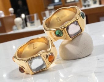 May include: Two gold-colored rings with a textured finish. Each ring features a square, light-colored stone and smaller, colorful gemstones. One ring has the number "8" and the other has the letter "L" engraved inside.