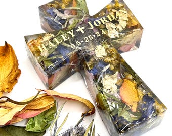 May include: A cross-shaped clear resin ornament filled with dried flowers in shades of yellow, blue, and green. The surface features gold lettering that reads HALEY + JORDAN and the date 05-25-24. Loose dried flower petals are scattered nearby.