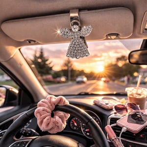 May include: A silver car visor clip featuring a rhinestone angel pendant. The angel has outstretched wings and a sparkling body. The interior of a car is visible, with a pink scrunchie on the steering wheel and a sunset in the background.