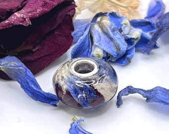 Op de afbeelding: Een ronde, heldere harskraal met ingebedde gedroogde blauwe en donkerpaarse bloemblaadjes. De kraal heeft een gepolijste zilverkleurige metalen kern met een breed middengat, ontworpen om aan een armband of ketting te worden geregen.