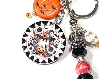 May include: A Halloween-themed keychain with a carved orange pumpkin charm and a round pendant featuring a ghost wearing a witch hat, surrounded by sunflowers and bats. The keychain includes decorative beads and a skull charm.