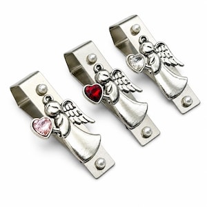 May include: Three silver-toned metal stocking holders, each featuring an angel figure holding a heart-shaped crystal. The angels are accented with small white faux pearls at the top and bottom of the rectangular metal clips, with heart crystals in light pink, deep red, and clear.