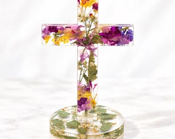 May include: A clear resin cross standing on a circular base, both containing embedded dried flowers in shades of purple, yellow, and white, along with green leaves. The cross features a vertical and horizontal bar design with visible floral textures.