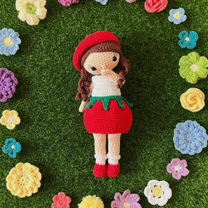 May include: A handmade crochet doll with a red beret, brown hair, and a white top. The doll has a red skirt with a green trim and red shoes. The doll is surrounded by colorful crochet flowers on a green background.