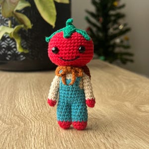 May include: A handmade crochet tomato doll with a smiling face, green leafy top, and blue overalls. The doll has a tan scarf and red accents on the feet and hands. The doll is approximately 4 inches tall.