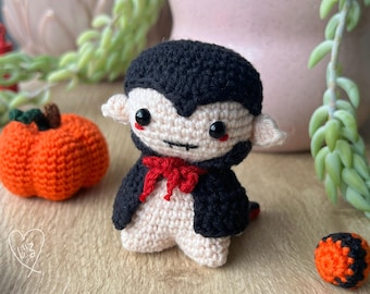 May include: A small crocheted vampire doll with a black cape and red bow tie. The doll has a white face with black eyes and a small mouth. The doll is sitting on a wooden surface with a small orange pumpkin and a small orange and black ball.
