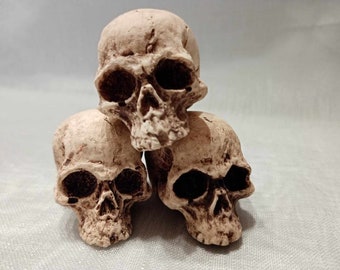 May include: Three realistic-looking white skull figurines stacked on top of each other. The skulls are made of a smooth, white material and have detailed features, including eye sockets, nasal cavities, and teeth.