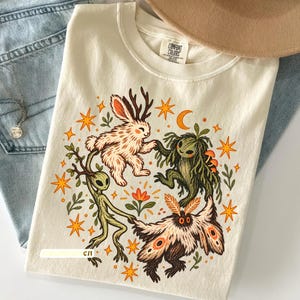 May include: A cream-colored short-sleeve t-shirt with a Comfort Colors label at the neck. The graphic features a circular illustration of a jackalope, a green forest creature, and a moth holding hands, surrounded by orange stars, a moon, and foliage.