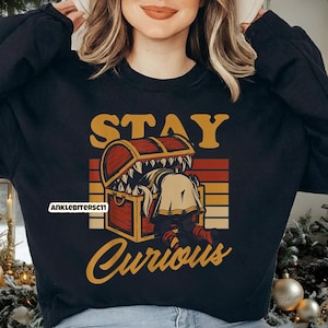 May include: Black sweatshirt with a graphic design featuring a treasure chest with a monster inside. The text "STAY Curious" is in a retro font. The design includes orange, yellow, and brown colors.