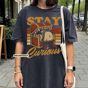 May include: Dark gray t-shirt with a graphic design featuring a treasure chest with a monster inside. The text "STAY Curious" is above the chest in orange and yellow. The shirt has a vintage, washed look.