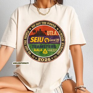 May include: Cream-colored t-shirt with a circular graphic featuring text and a cityscape silhouette. The graphic includes the words "WE GOT THE POWER," "UTLA," "SEIU Local 99," "TEAMSTERS AALA Local 2010," and "2026."
