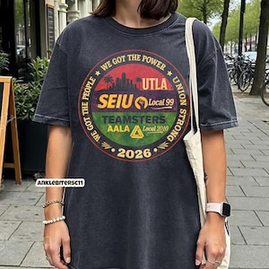 May include: Dark gray t-shirt with a circular graphic featuring the text "WE GOT THE POWER" and "UNION STRONG". The graphic includes the text "UTLA", "SEIU Local 99", "TEAMSTERS AALA Local 2010", and the year "2026".