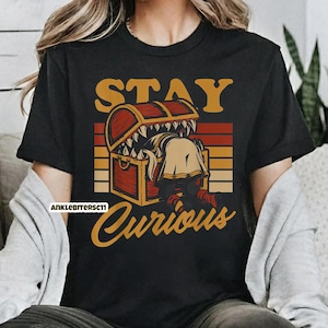 May include: Black t-shirt with a graphic featuring a treasure chest with teeth and a person reaching inside. The text "STAY CURIOUS" is above and below the chest in a vintage style.