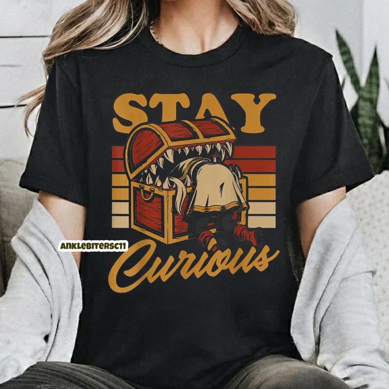May include: Black t-shirt with a graphic featuring a treasure chest with teeth and a person reaching inside. The text "STAY CURIOUS" is above and below the chest in a vintage style.