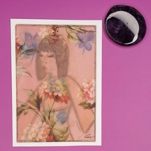 May include: A framed artwork with a pink background features a woman's portrait surrounded by floral elements in shades of pink, white, and purple. A dark, oval-shaped stone with a white swirl is positioned to the right of the artwork.