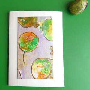 Puede incluir: Una tarjeta de felicitación blanca con una pintura abstracta de círculos verdes y naranjas sobre un fondo lavanda. La tarjeta está sobre una superficie verde, con una piedra verde y amarilla en la esquina superior derecha.