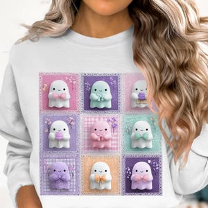 Pode incluir: Camisola branca com um design de patchwork de fantasmas coloridos em tons pastel. Cada fantasma segura um laço, uma flor ou um ovo de Páscoa. Os quadrados têm uma borda de renda.