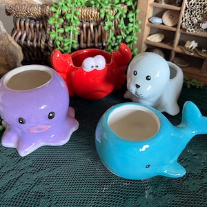 May include: A collection of ceramic planters in the shape of sea creatures. Includes a purple octopus, a red crab, a blue whale, and a light blue seal. Each planter has a white interior and is designed with cartoon-like features.