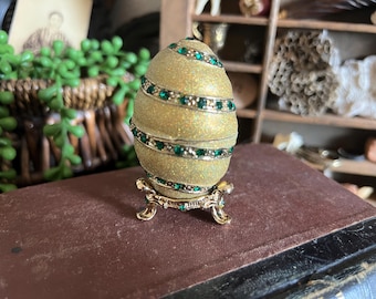 Könnte beinhalten: Ein dekoratives, goldfarbenes, eiförmiges Ornament mit smaragdgrünen Akzenten. Das Ei steht auf einem goldfarbenen Ständer und ruht auf einer braunen Oberfläche. Das Ei hat ein Spiraldesign mit eingebetteten grünen Steinen, was einen Hauch von Eleganz verleiht.