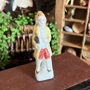 May include: A porcelain figurine of a man wearing a yellow jacket, red pants, and a white shirt. The figurine is standing on a white base.