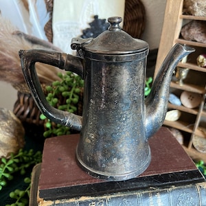 May include: A silver teapot with a tarnished finish. The teapot has a rounded body and a spout on the side. It is sitting on a stack of books.