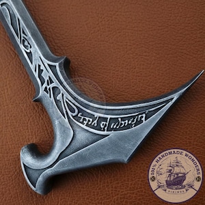 May include: A detailed close-up of a silver-colored, curved dagger with intricate designs and engraved text. The blade has a sharp point and a decorative handle. The image includes a circular logo with a ship and the text "Handmade Wonders Vikings".