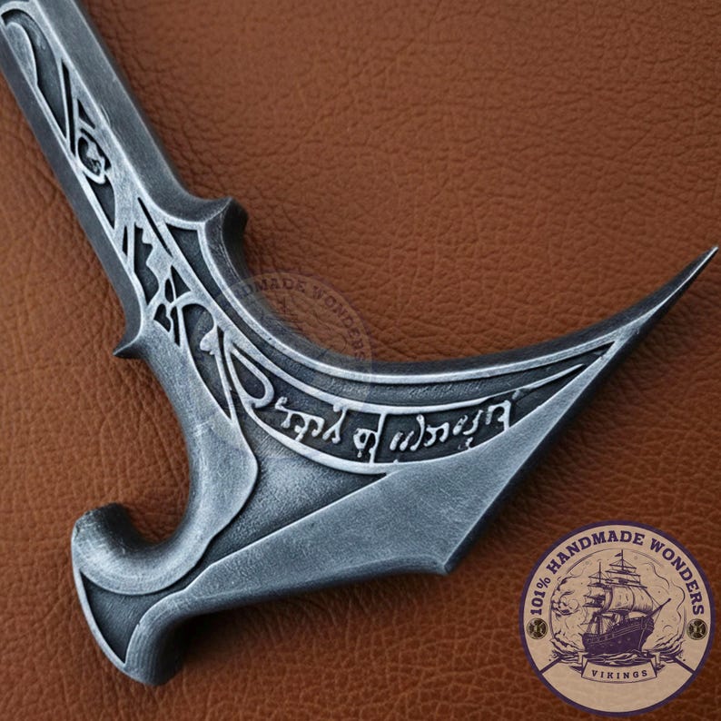 May include: A detailed close-up of a silver-colored, curved dagger with intricate designs and engraved text. The blade has a sharp point and a decorative handle. The image includes a circular logo with a ship and the text "Handmade Wonders Vikings".