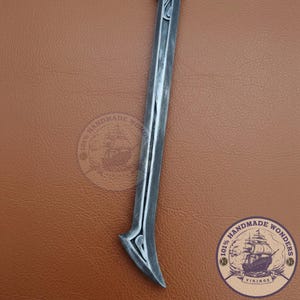 May include: A silver-colored, curved blade with a decorative handle. The blade has a central groove and a pointed tip. The handle features an ornate design. The background is a brown leather-like surface.