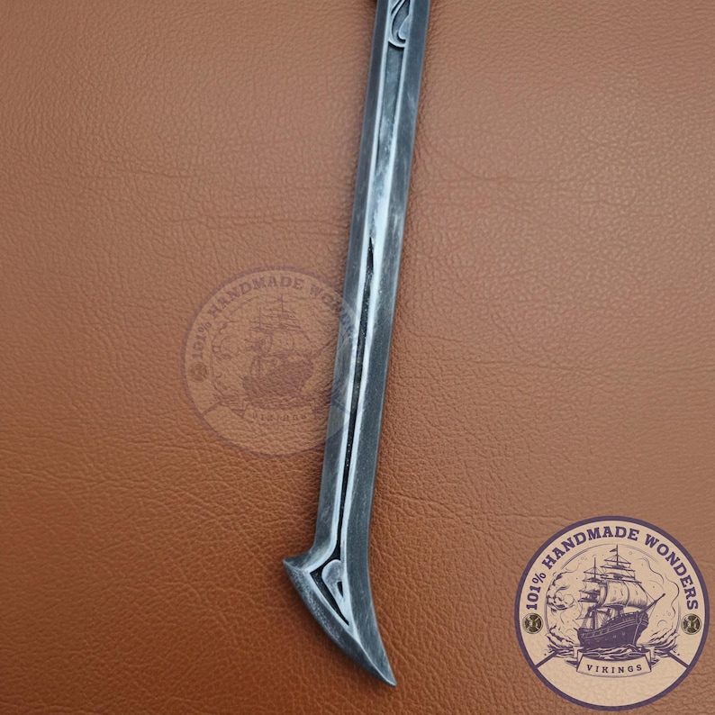 May include: A silver-colored, curved blade with a decorative handle. The blade has a central groove and a pointed tip. The handle features an ornate design. The background is a brown leather-like surface.
