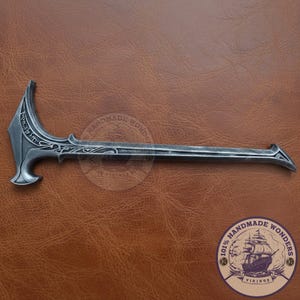 May include: A silver-colored, ornate, Viking-style weapon with a curved blade and a long handle. The weapon features intricate carvings and a decorative design. The handle has a pointed end. The weapon is displayed on a brown leather surface.