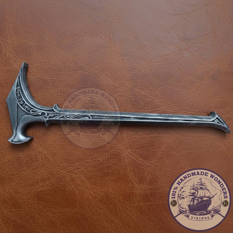 May include: A silver-colored, ornate, Viking-style weapon with a curved blade and a long handle. The weapon features intricate carvings and a decorative design. The handle has a pointed end. The weapon is displayed on a brown leather surface.