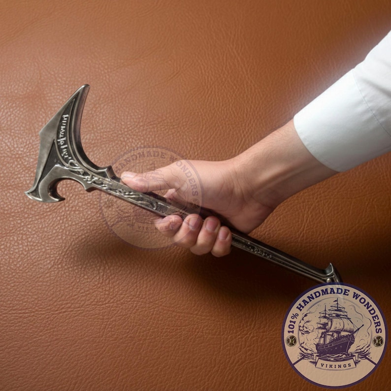 May include: A silver-colored, ornate axe with a curved blade and detailed handle. The axe is held in a hand against a brown leather background. The axe has decorative carvings and a pointed end.