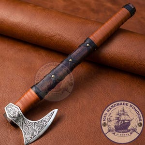 Könnte beinhalten: Eine handgefertigte Axt mit einer silbernen Klinge mit aufwendigen Designs. Der Griff ist mit braunem Leder mit schwarzen Akzenten und Messingnieten umwickelt. Die Axt wird auf einer braunen Lederoberfläche präsentiert.