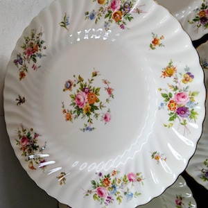 May include: White porcelain plate with a floral pattern of pink, orange, yellow, and purple flowers. The plate has a scalloped edge and a gold trim. The floral arrangement is clustered in the centre and around the rim.
