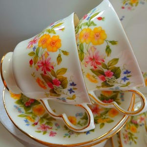 Op de afbeelding: Twee witte theekopjes en schotels met een bloemenpatroon. De kopjes en schotels hebben een gouden rand en zijn versierd met gele, roze en blauwe bloemen en groene bladeren. De theekopjes zijn op elkaar gestapeld.
