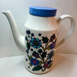 May include: A white ceramic teapot with a blue lid and a curved spout. The teapot is decorated with a floral pattern in shades of blue, pink, and green. It has a large handle for easy pouring.