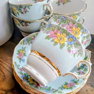 May include: A collection of vintage teacups and saucers. The cups and saucers are pale blue with a floral design in pink, yellow, and blue. The rims and handles are gold. The set includes six cups and saucers.