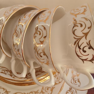 May include: A set of white porcelain teacups and saucers with gold filigree detailing. The cups are stacked, showcasing their curved handles and the intricate gold patterns along the rims and surfaces. The saucers below mirror the design.