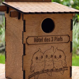 May include: A wooden birdhouse with a brown stain finish. The front of the birdhouse has a sign that reads "Hôtel des 3 Piafs" and a laser-engraved image of three birds perched on a branch.