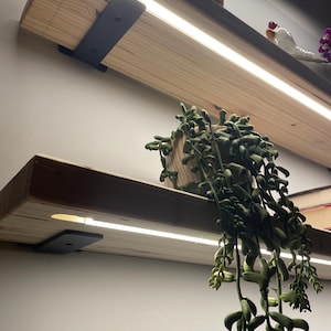 May include: Two light-coloured wooden floating shelves mounted with black metal brackets. Each shelf features a long, integrated LED light strip embedded along the front edge, casting a bright white glow downwards onto the space below.