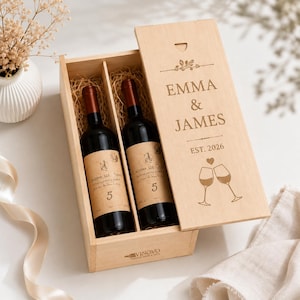 Könnte beinhalten: Eine Weinkiste aus hellem Holz mit zwei dunklen Glasweinflaschen, die in Stroh gebettet sind. Der Schiebedeckel ist mit dem Text EMMA & JAMES, EST. 2026 und einer Abbildung von zwei anstoßenden Weingläsern mit einem kleinen Herz graviert.