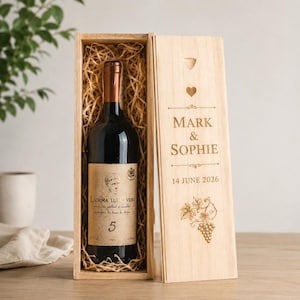 Puede incluir: Una botella de vino de vidrio oscuro con etiqueta beige que dice Lacrima lui Ovidiu 5, dentro de una caja de madera clara con paja. La tapa de la caja tiene grabado el texto Mark & Sophie, la fecha 14 de junio de 2026, un corazón y una vid.