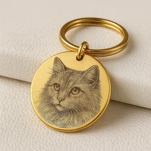 Può includere: Un portachiavi rotondo color oro con un ritratto inciso dettagliato di un gatto. Il pelo del gatto è raffigurato con linee sottili. Il portachiavi ha un anello diviso per attaccare le chiavi.