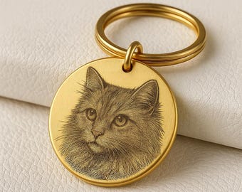 May include: A gold-colored round keychain with a detailed engraved portrait of a cat. The cat has long fur and is facing forward. The keychain ring is also gold-colored and attached to the top of the pendant.