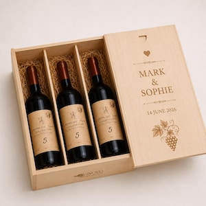 Könnte beinhalten: Eine Weinkiste aus hellem Holz mit drei Flaschen Rotwein, die in Stroh gebettet sind. Der Schiebedeckel ist mit einem Herz, den Namen MARK & SOPHIE, dem Datum 14 JUNE 2026 und einer Weintrauben-Illustration graviert. Die Marke VINOVO ist sichtbar.