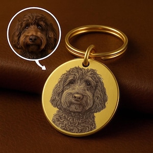 Puede incluir: Un llavero redondo de color dorado con una imagen grabada detallada de la cara de un perro. El perro tiene pelo rizado de color marrón. El llavero tiene un anillo dorado para sujetar las llaves.