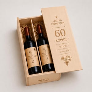 May include: A light-coloured wooden wine box containing two dark glass wine bottles nestled in straw. The sliding lid is engraved with AGED TO PERFECTION, 60, SOPHIE, EST. 1966, 14 JUNE 2026, and a grape vine illustration. The brand VINOVO is on the box base.