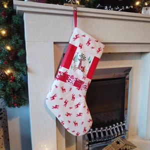 May include: A white and red Christmas stocking with a patchwork design featuring reindeer and a Christmas tree. The stocking is hanging from a hook above a fireplace.