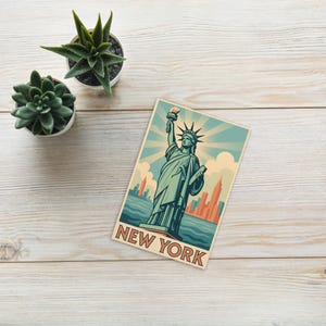 Könnte beinhalten: Eine Postkarte im Vintage-Stil mit der Freiheitsstatue in Türkis- und Beigetönen und dem Text "NEW YORK" am unteren Rand. Der Hintergrund zeigt eine Skyline und Sonnenstrahlen. Zwei kleine Sukkulenten in Töpfen sind zu sehen.